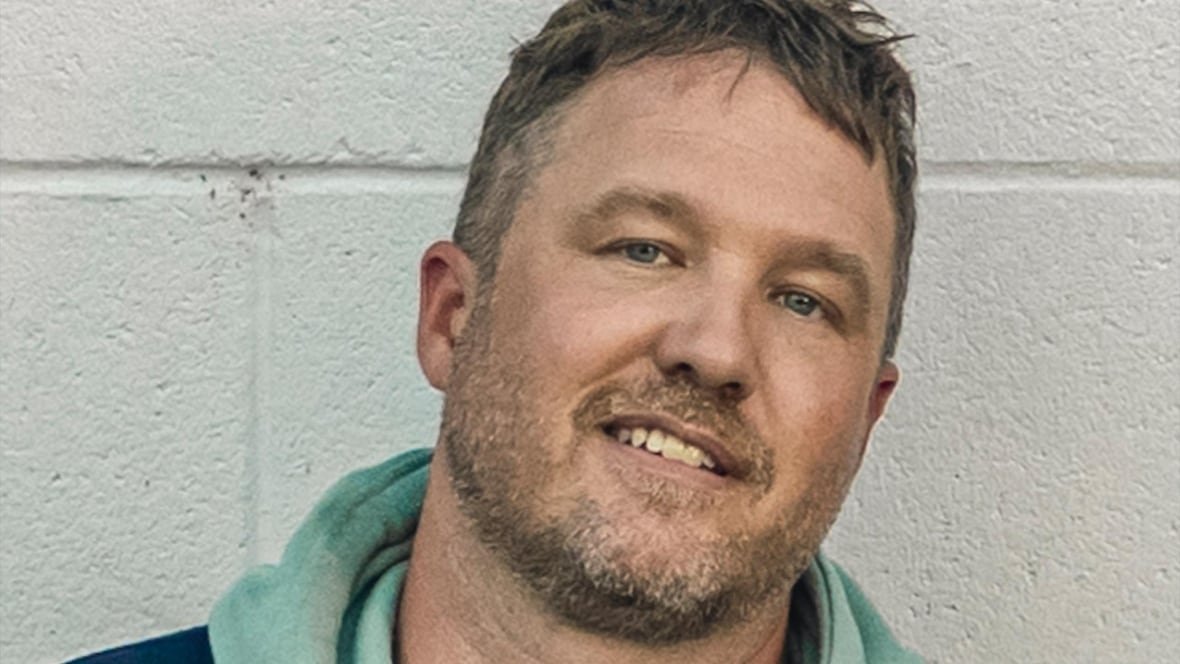 Greg, a white man with brown hair and blue eyes, has brown facial hair and wears a green hoodie while standing in front of a white wall.