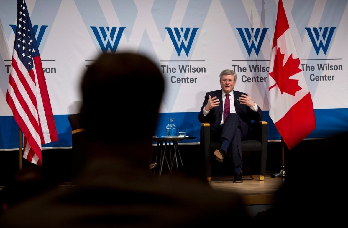 harper-washington-20120402-1 A man on stage gestures while addressing someone, seen in silhouette, in the audience. There are U.S. and Canadian flags on stage.