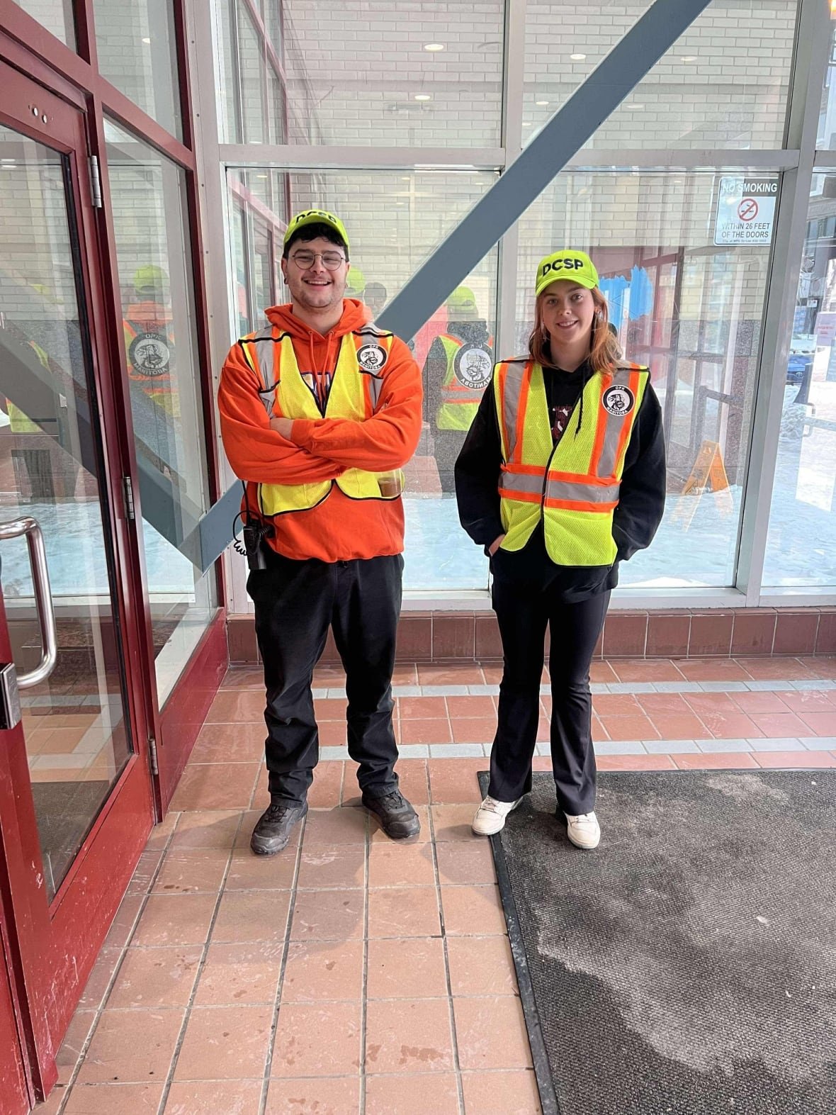 helpers-justin-bourbonniere-and-ashley-hunter Two young people in reflective vests stand in front a large window at the door ways of a shopping centre.