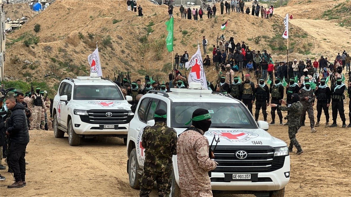 Two vans that appear to have a Red Cross symbol on them are shown travelling on a dirt path. Around them, and in the distance, are armed men, some in camouflage and with faces covered.