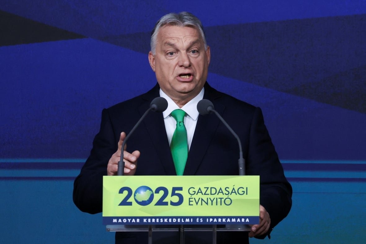 A cleanshaven grey-haired man wearing a suit and tie speaks in front of a podium and a microphone.
