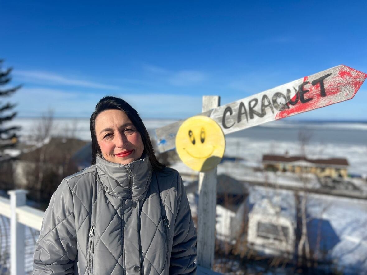 A woman poses for a photo in front of a Caraquet sign with an Acadian flag motif