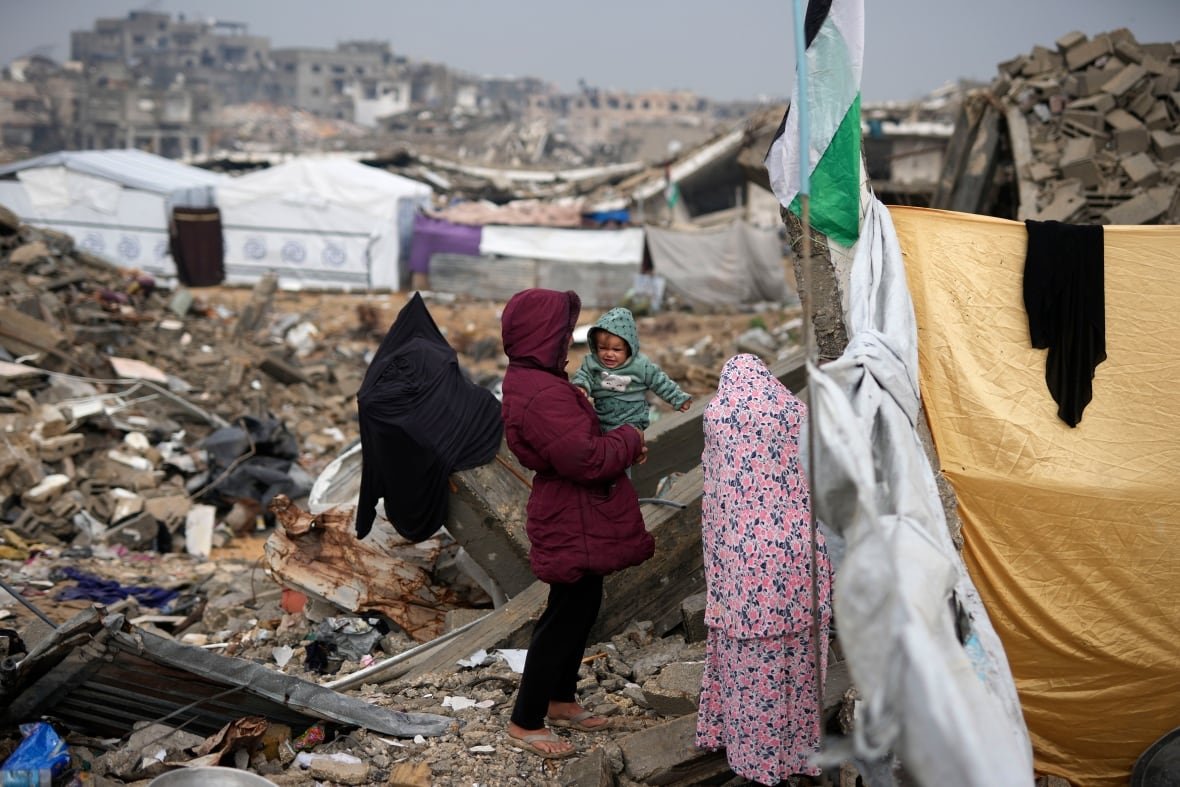 israel-palestinians-gaza In the centre of the image, a person in a maroon parka is holding a baby. The baby seems upset. They are standing outside of a tent with piles of rubble surrounding them on all sides and more tents visible in the distance.