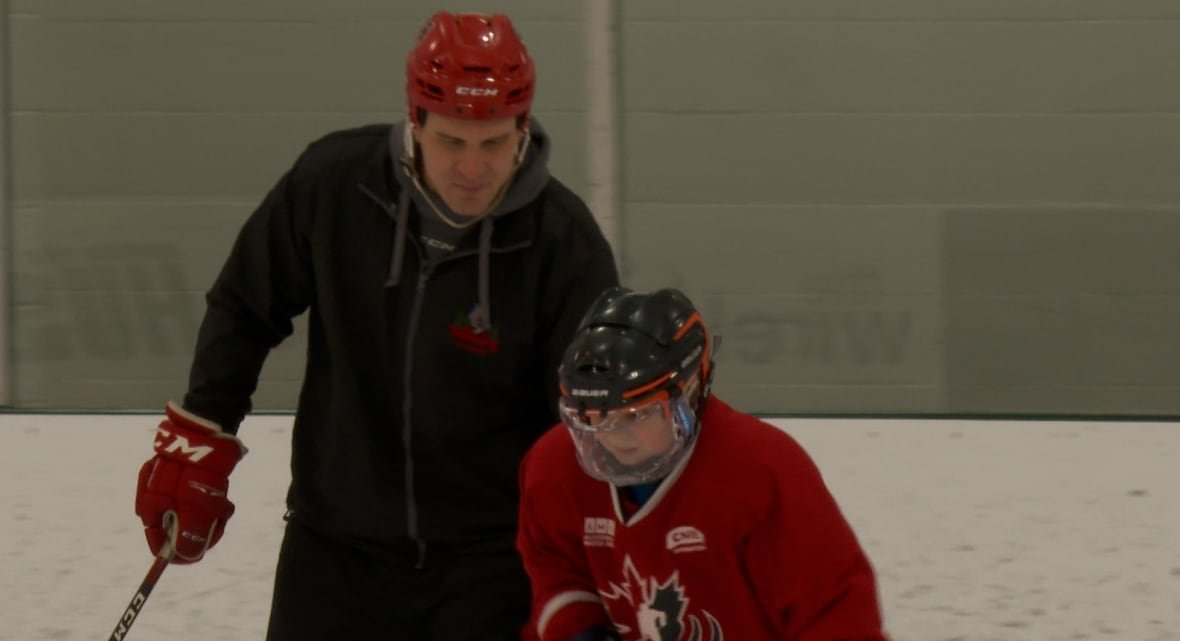 An adult helps a young person in a red jersey and hockey equipment.