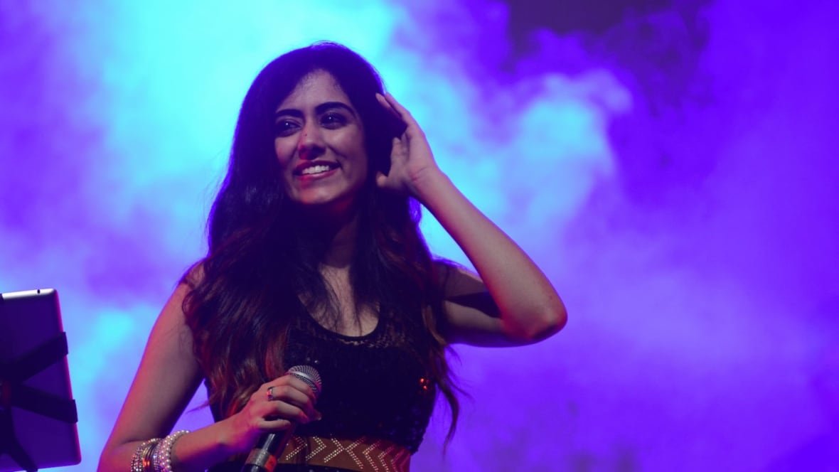 jonita-gandhi A smiling woman holding a microphone stands in front of a wall of smoke.