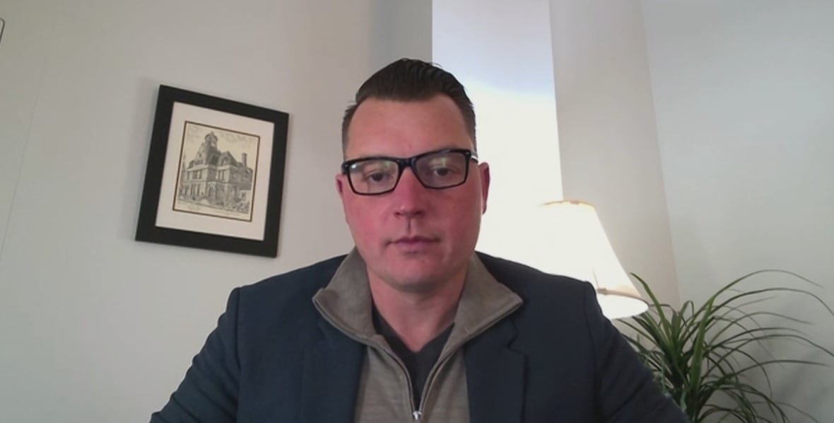 Man wears sweater, suit jacket and glasses as he looks into camera.