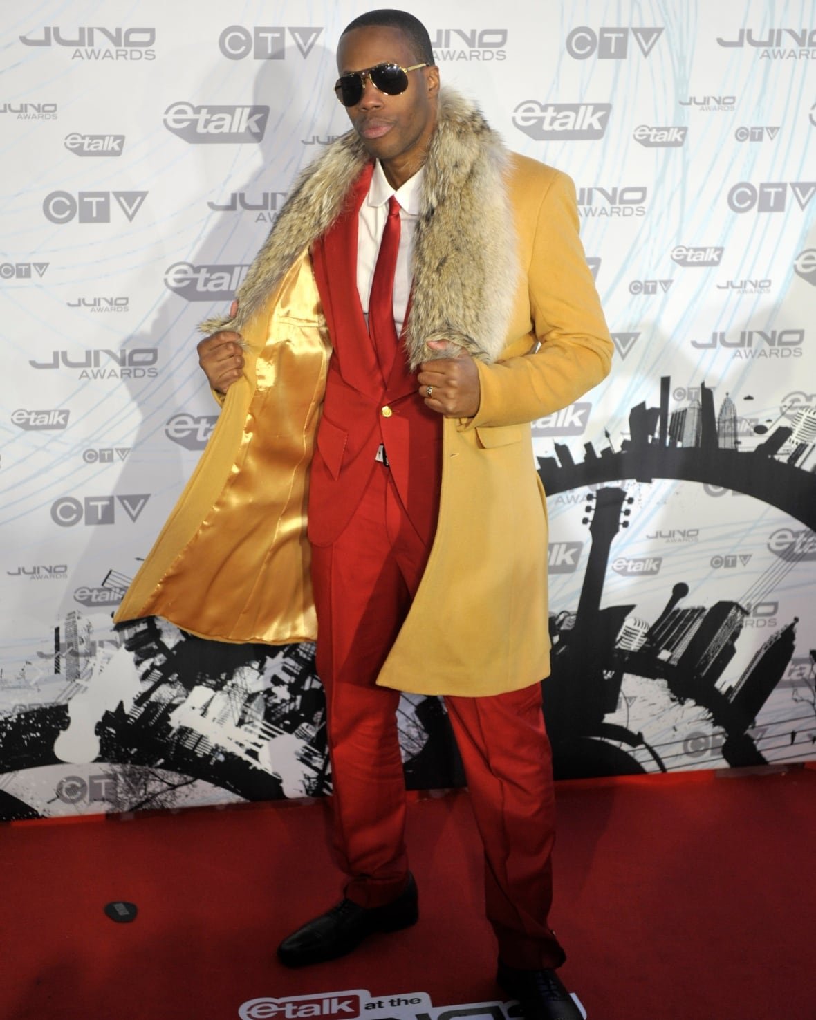Kardinal Offishall on the red carpet wearing a red suit and tie with a gold-toned, fur-trimmed wool coat.