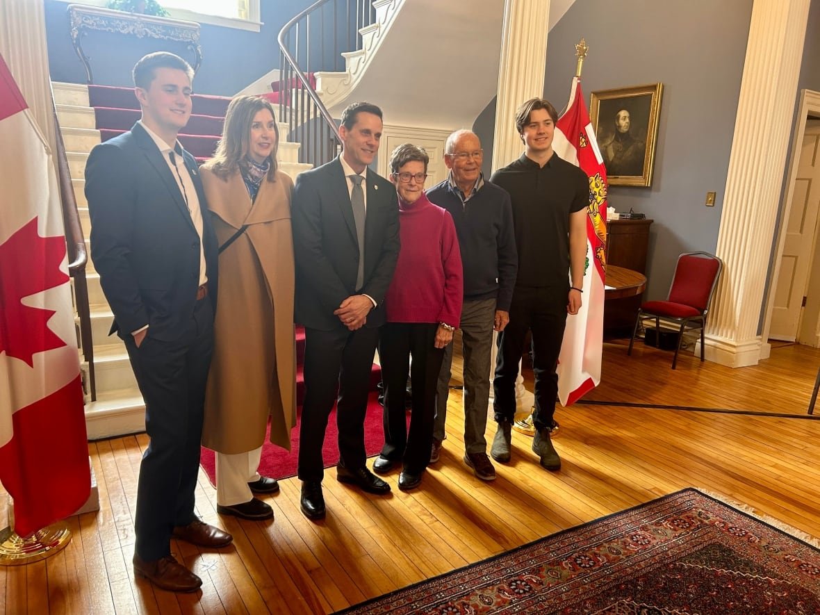 Six people stand at the bottom of an ornate staircase, backlit by the sun coming through a window: Two young men, a woman with long brown hair, a man in a business suit, and an older man and woman