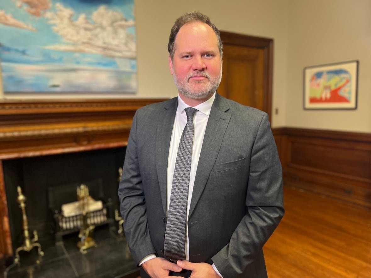 manitoba-justice-minister-matt-wiebe A man wearing a great suit and tie stands in an office with a fireplace and wooden floors in the background, and framed art on the walls.