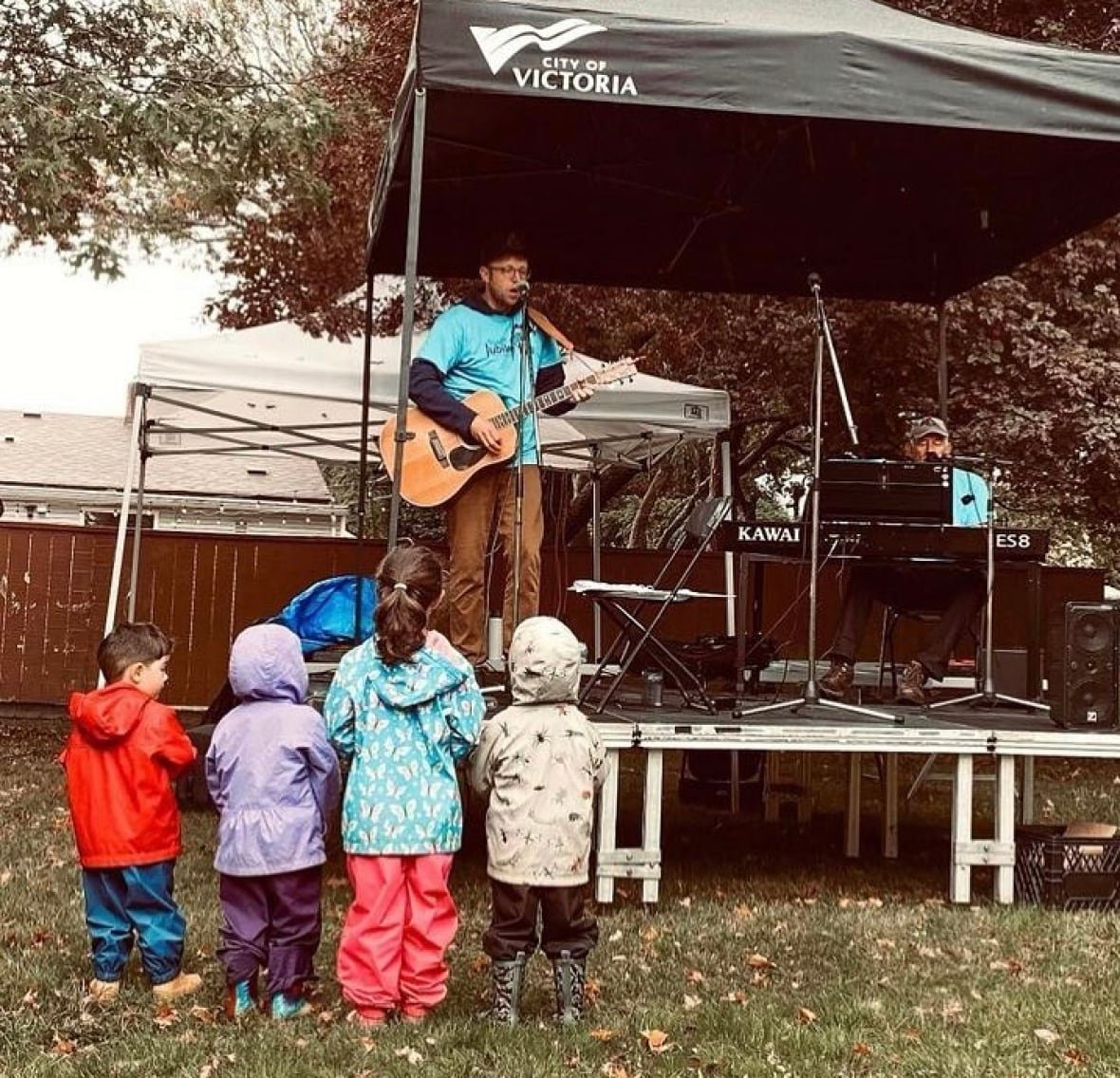 A man plays guitar and sings on stage as four small children, wearing rain jackets, look up at him.