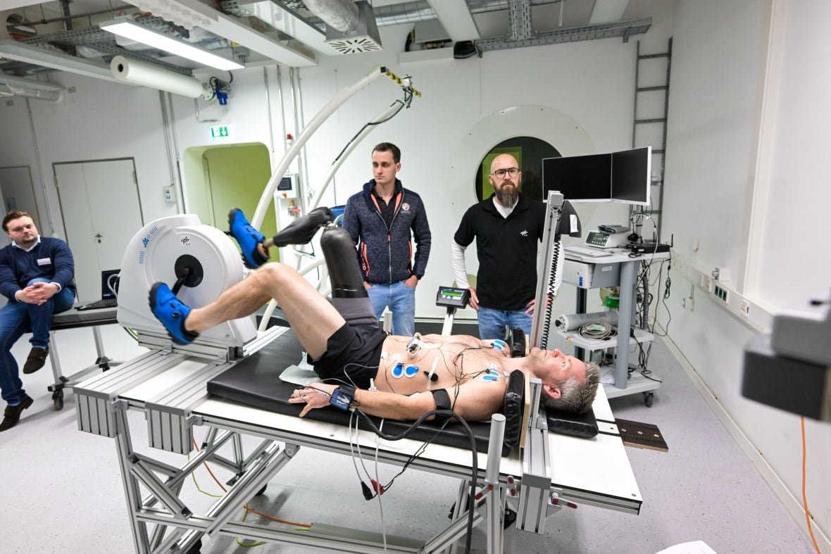 A man with a prosthetic leg lays back on a recumbent bike, pedalling. Three other men stand around and watch.