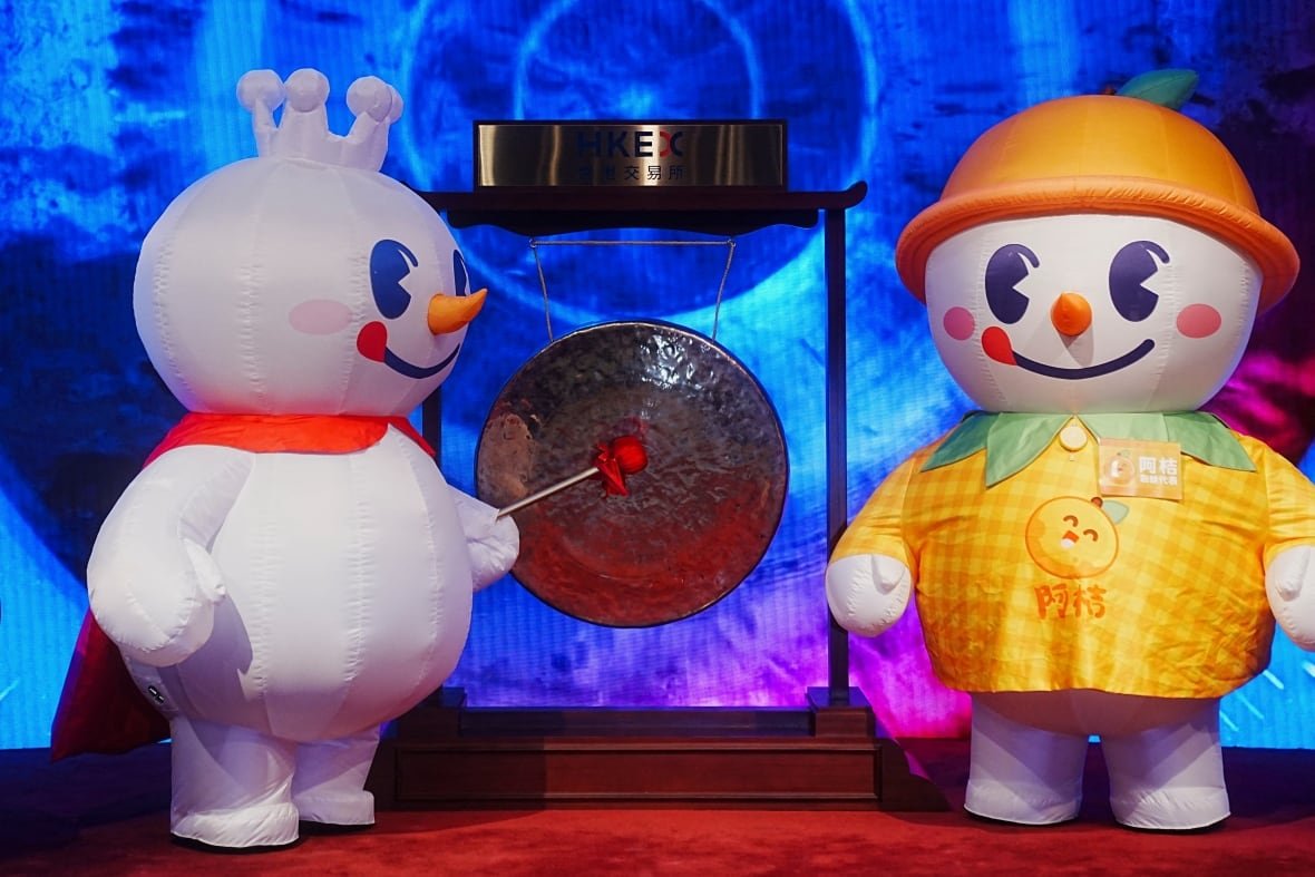 mixue-ipo A person in an inflatable snow man mascot suit strikes a gong on a stage as another snow man mascot character looks on.