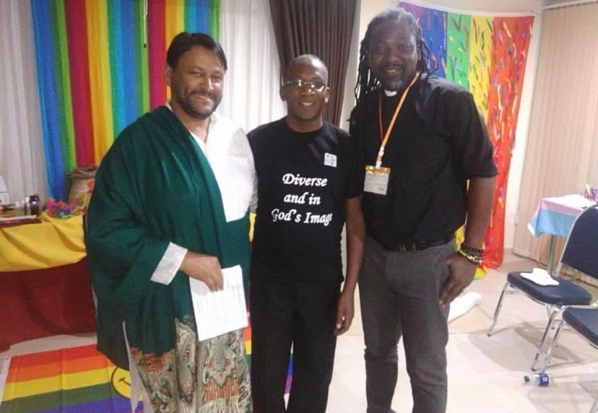 muhsin-hendricks-and-jide-macaulay Three men stand side-by-side smiling in a room decorated with rainbows. The man on the left wears long white robes with a green piece of fabric draped over his shoulder. The man on the right is wearing a black reverend's tunic. The man in the middle is wearing a T-shirt that reads: "Diverse and in God's image."
