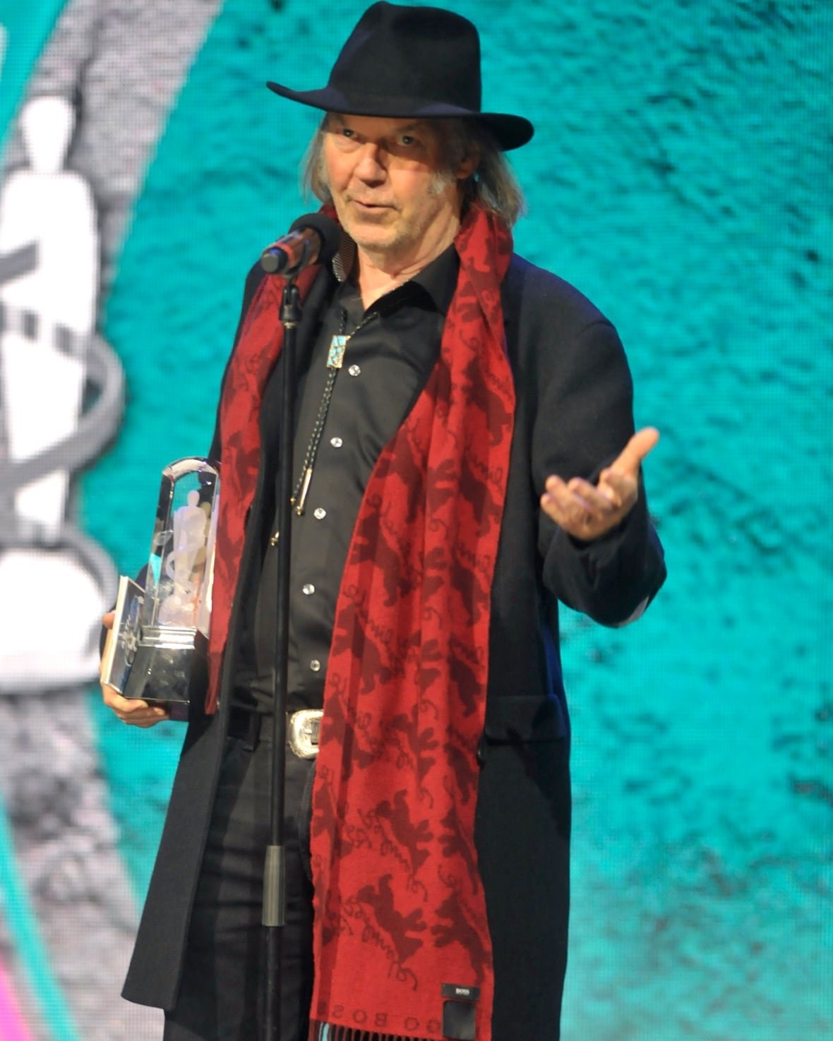 Neil Young on stage wearing a black coat, black dress shirt, black jeans, bolo tie and cowboy hat.