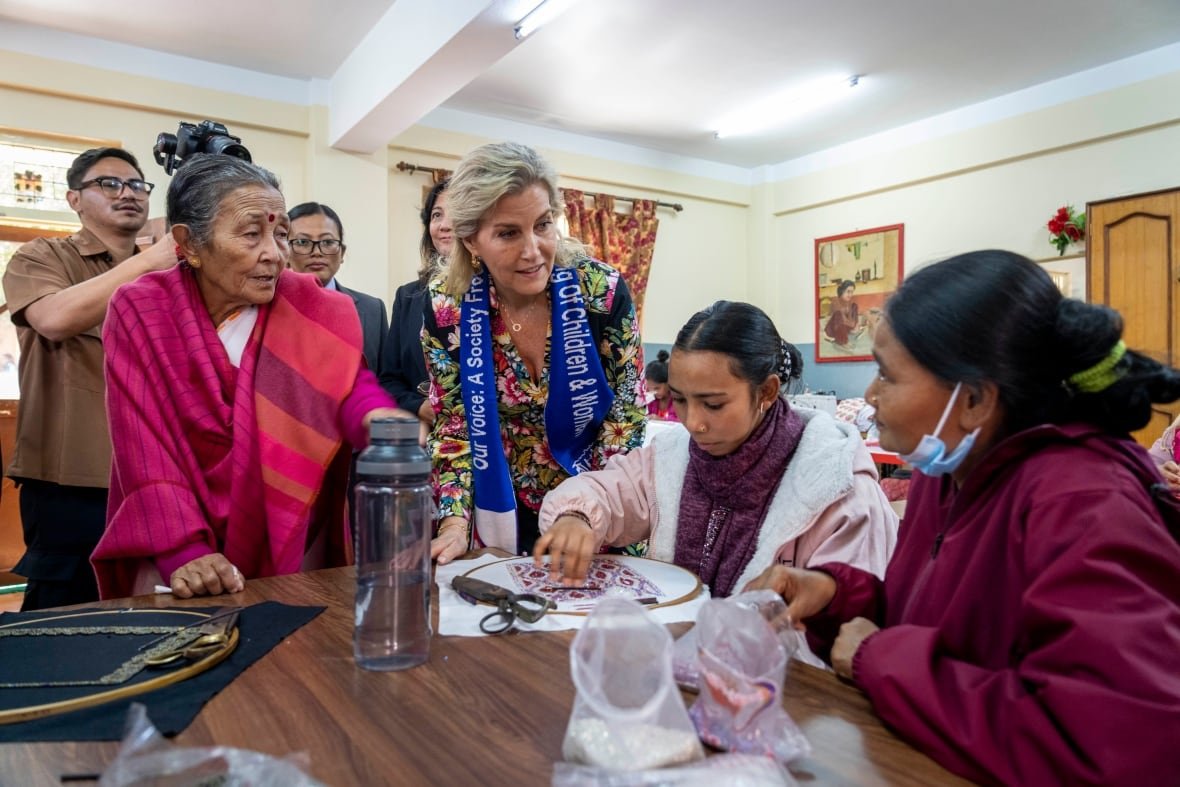 nepal-britain-royals A person sits at a table doing needlework as a person sitting nearby looks along. People standing nearby also look on.