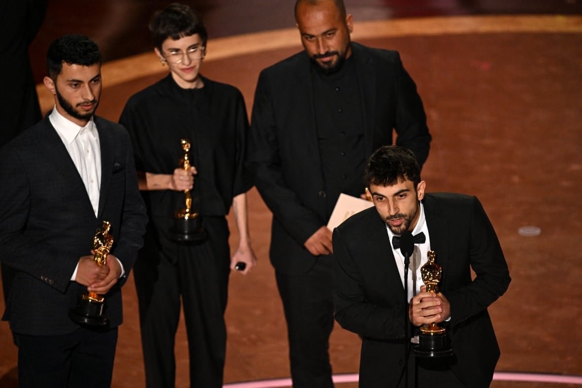 no-other-land A man speaks into a microphone with three people watching behind him.