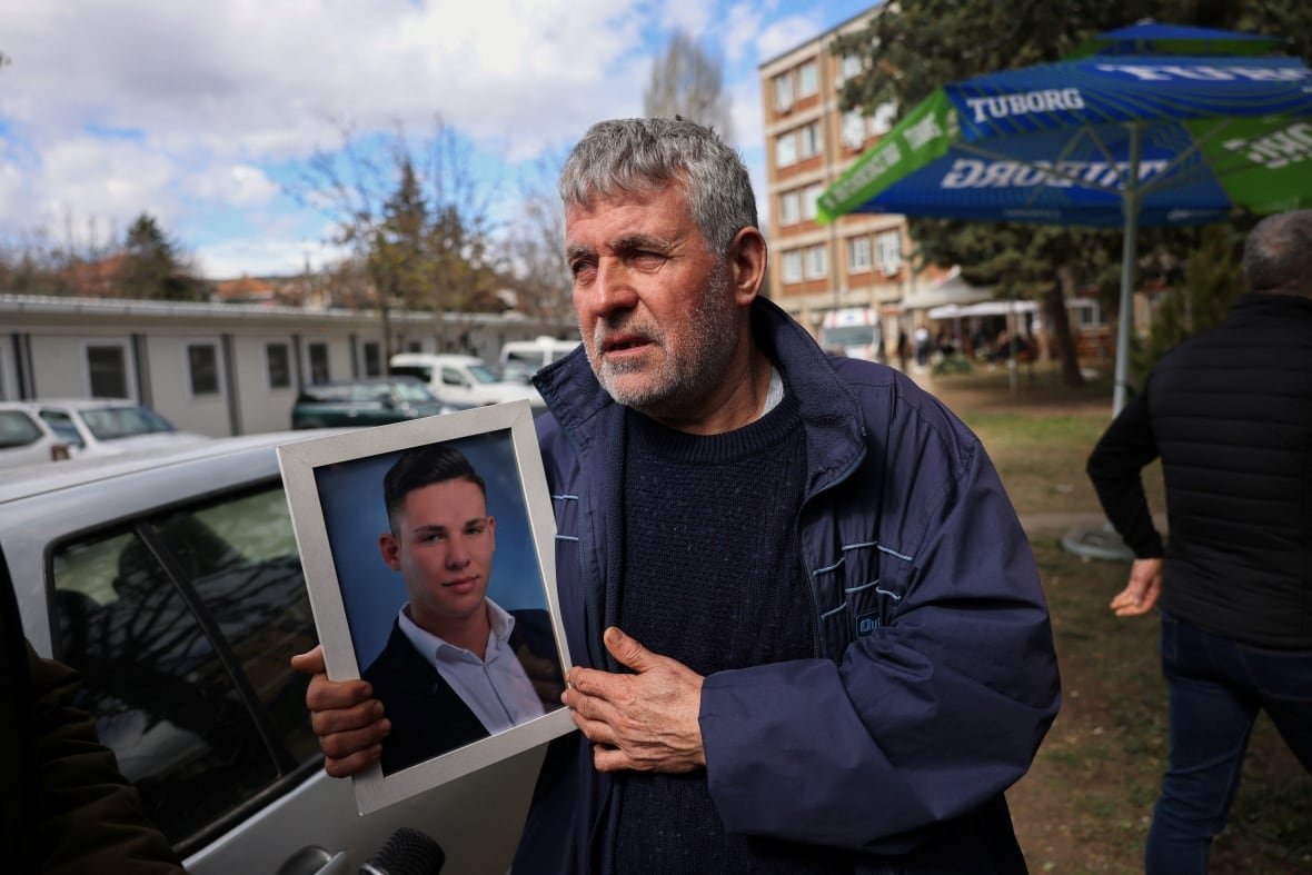 north-macedonia-fire-1 A bearded, grey-haired man in a coat outdoors holds up a framed photo of a younger man.