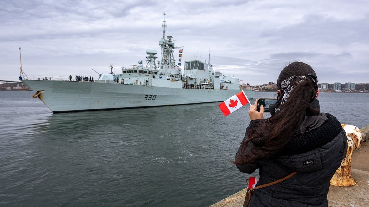 ns-nato-ship-20220319 A warship is seen in a body of water with a person waving a Canadian flag on the shore.