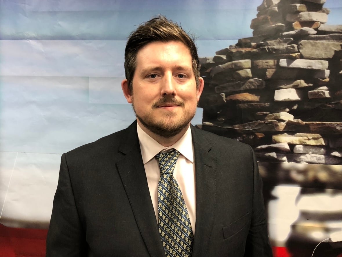 A man in a suit stands in front of a picture of an inukshuk.