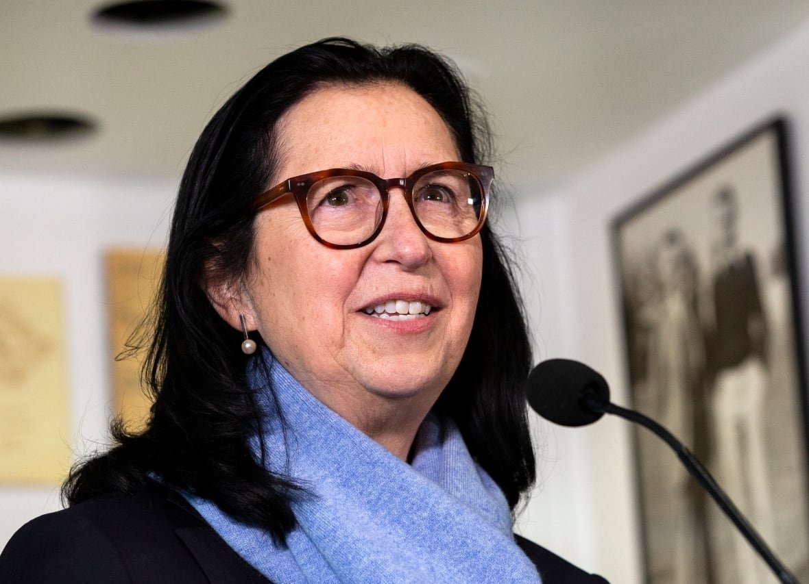 oly-canada-williams-medals-20230224 A woman wearing glasses and a scarf speaks into a microphone at a podium during a press conference.