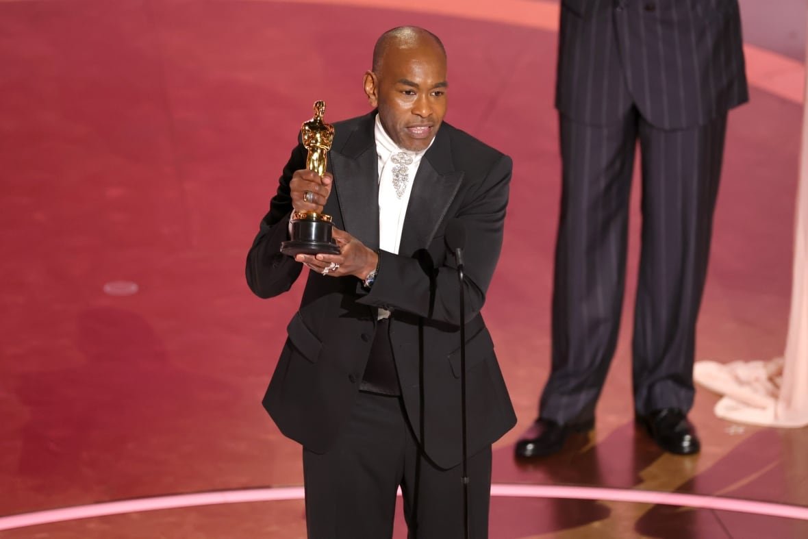 paul-tazewell A man wearing a black suit holds a trophy on a stage.