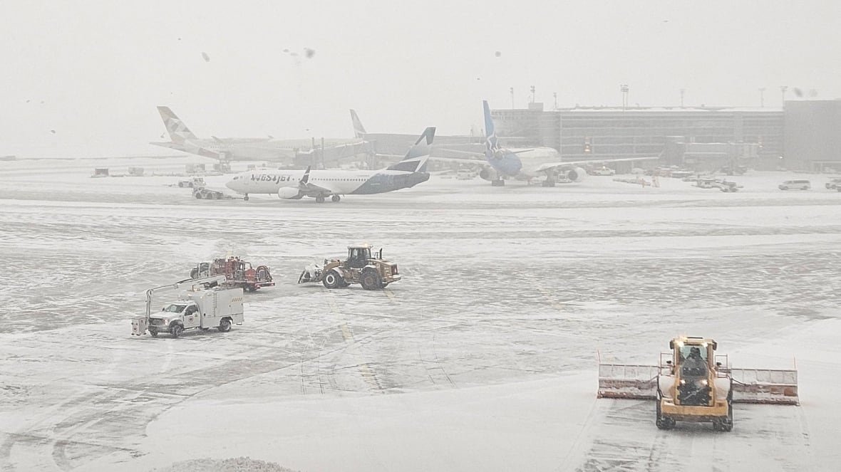 A near whiteout on an airport runway. Three planes are docked at the airport. Crews are clearing snow from the tarmac with machines