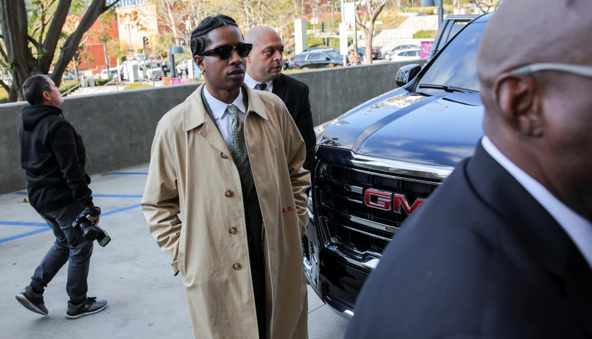 A man in a beige coat and sunglasses walks outside around the front of a car. Another man is following closely behind him.