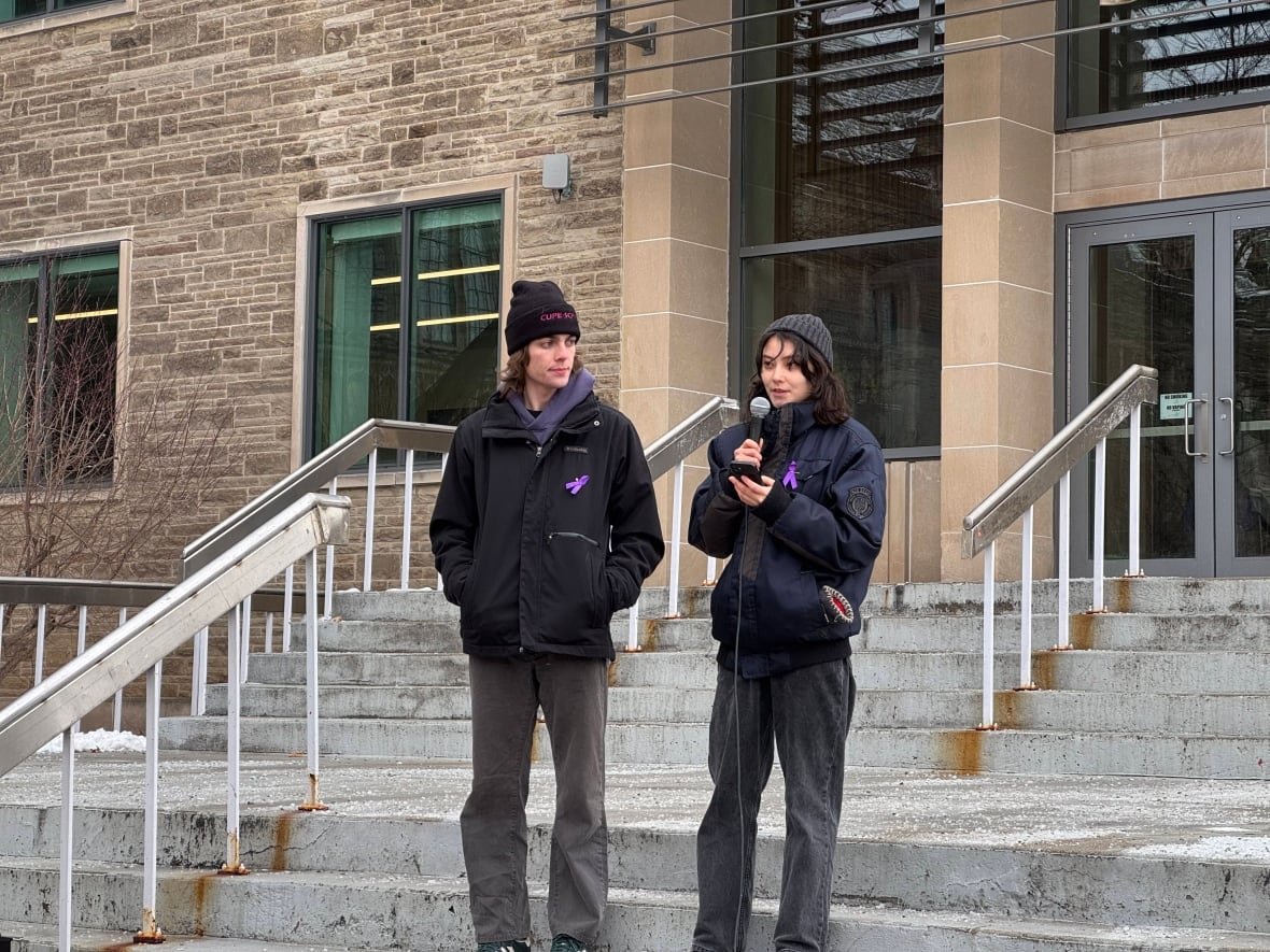 rally-at-mcmaster-1 Two people stand near the bottom of a stairway. One of them is speaking while holding a phone and a microphone.