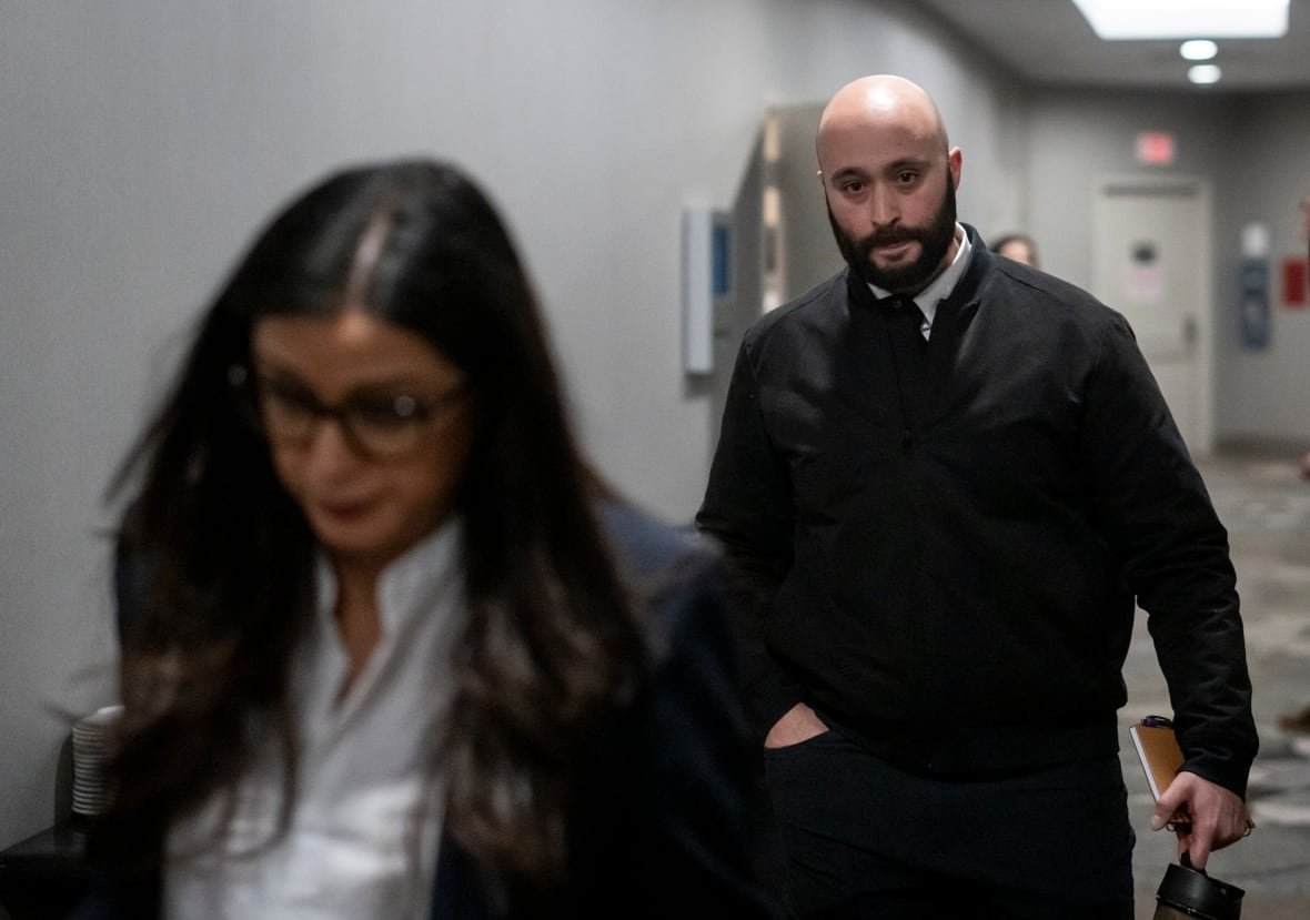 A man in a dark jacket, white shirt and dark tie walks behind a woman on his way out of a hearing.