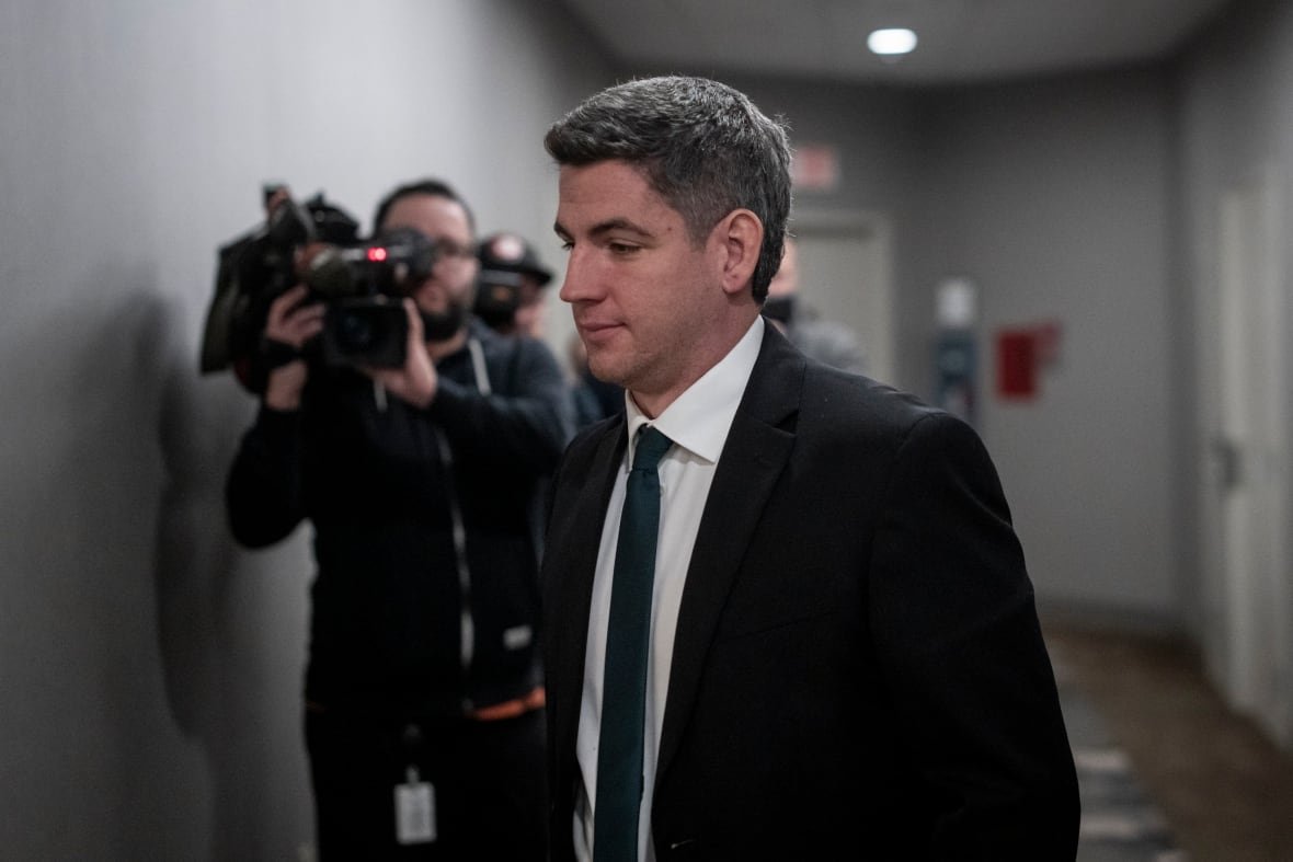 A man in a dark suit and tie walks part reporters with television cameras filming him.