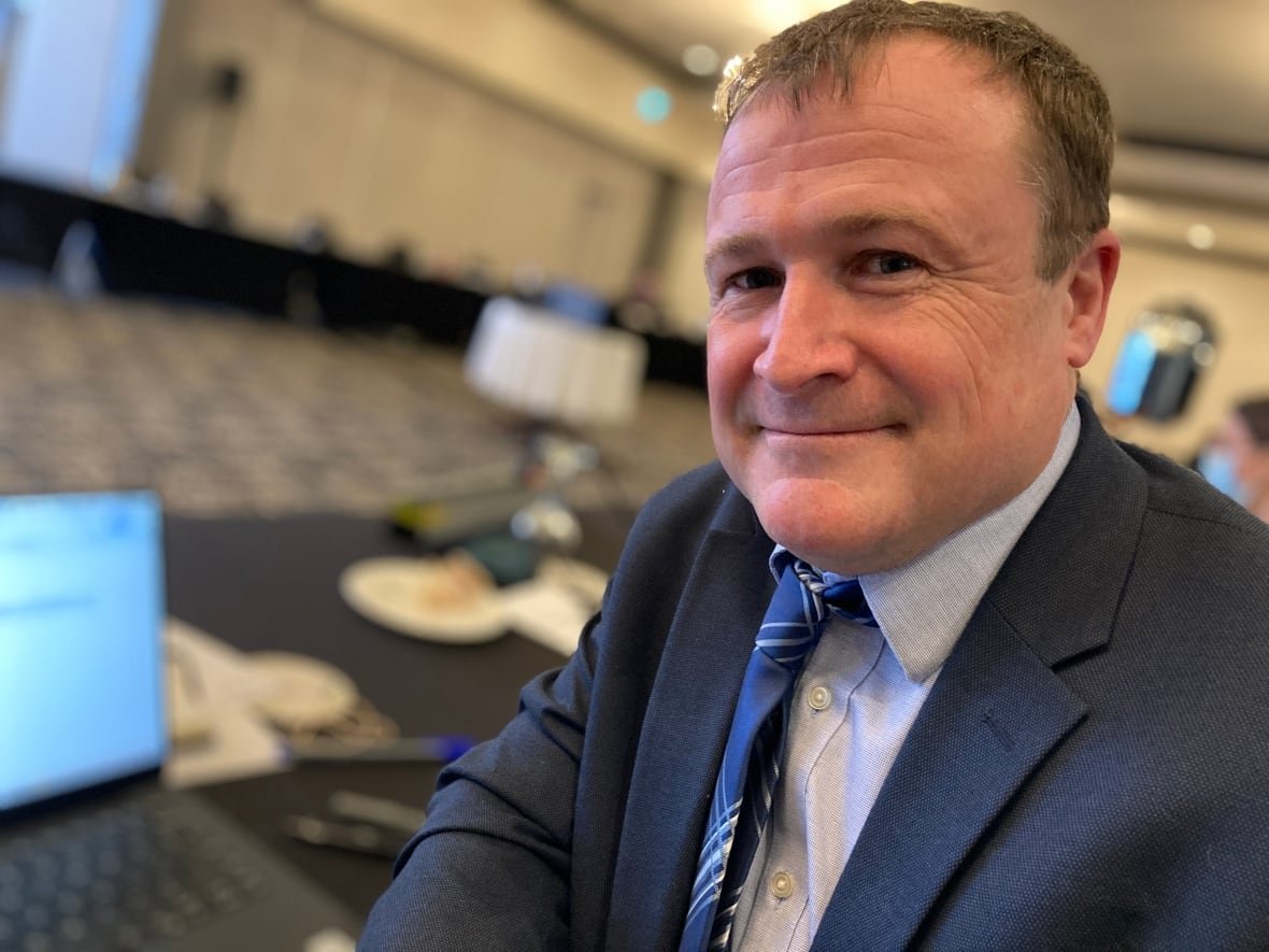 richard-saillant A man in a dark blue suit and a blue tie sits at a computer terminal, facing the camera with a smile.