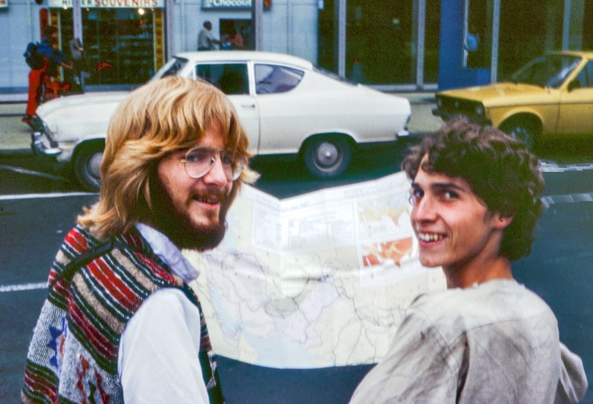 two young men in glasses hold a paper map. in the background is a street with cars parked on the side, the cars and their outfits are from the 1970s