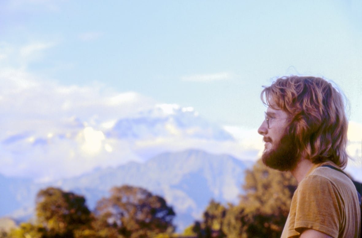 a man with shaggy hair and glasses looks out over the landscape. in the background trees and high mountain peaks are visible