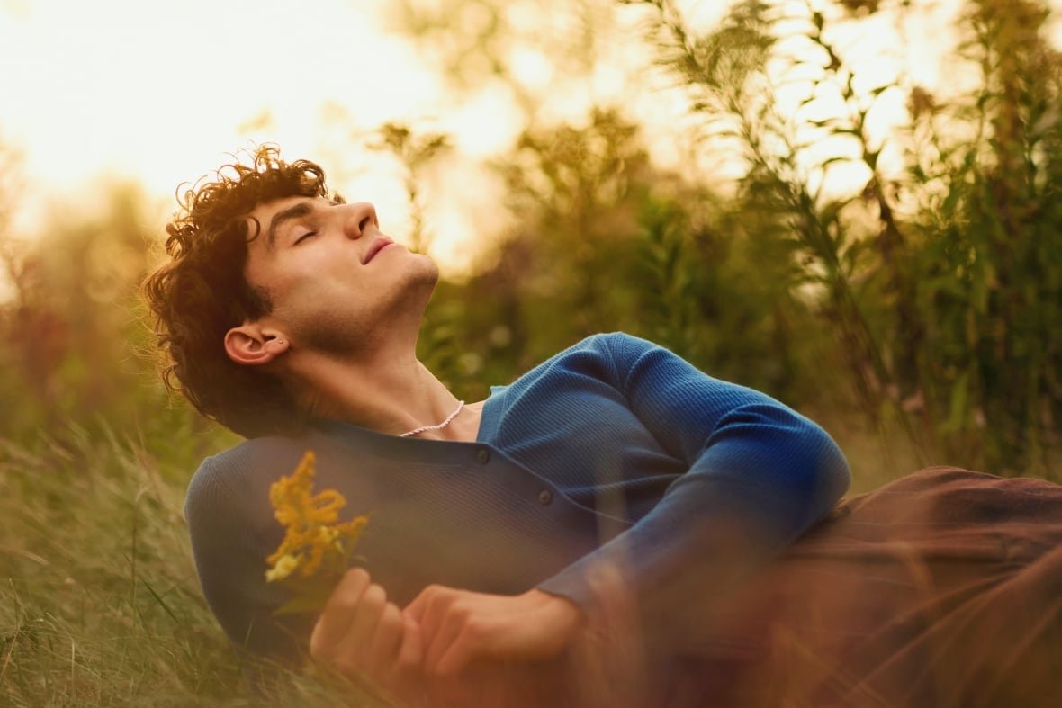 A young man with curly, dark hair wearing a blue, long-sleeved shirt and brown pants reclines in a field with his eyes closed while holding a wildflower.