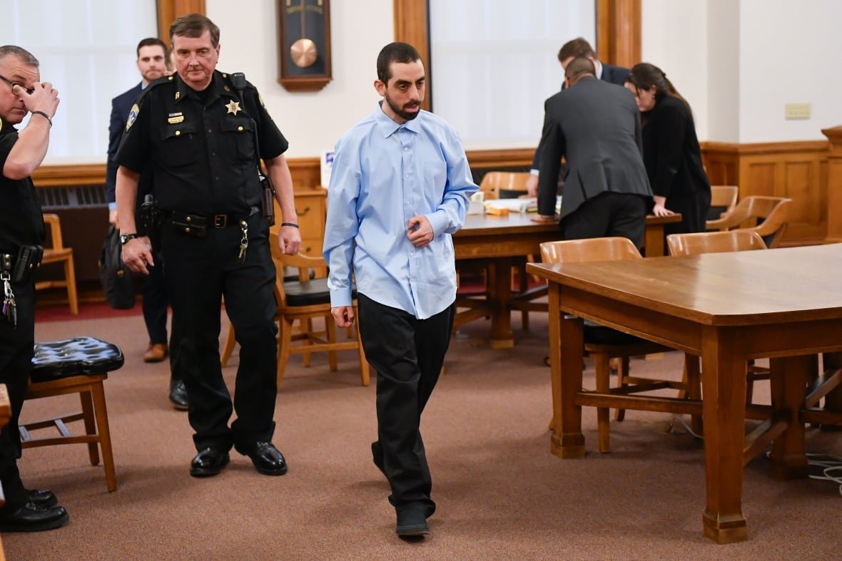 A man in a blue dress shirt walks in a courtroom, followed by police officers.
