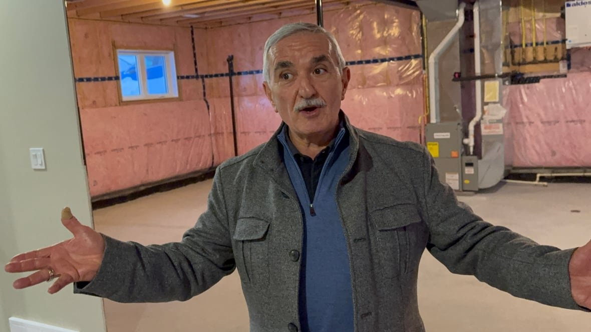 A man with grey hair and a mustache holds his arms wide in an unfinished basement