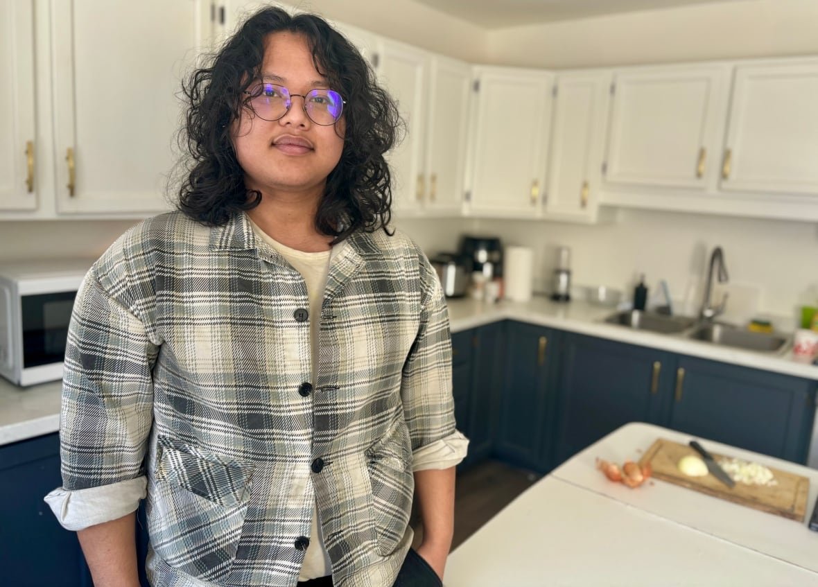 Man with glasses and long curly brown hair wears a grey and white plaid shirt as he stands in a kitchen.