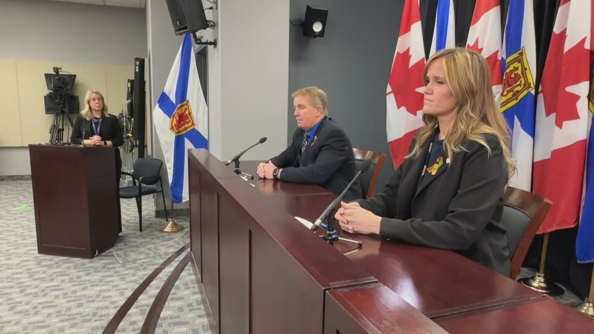 scott-armstrong-and-leah-martin A man and a woman sit at a podium with a backdrop of Canadian and Nova Scotian flags. A moderator stands to their left.