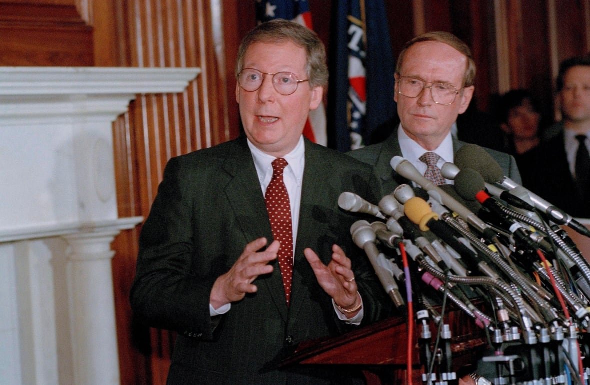 Two clean-shaven men wearing suit and tie stand near a bank of microphones.