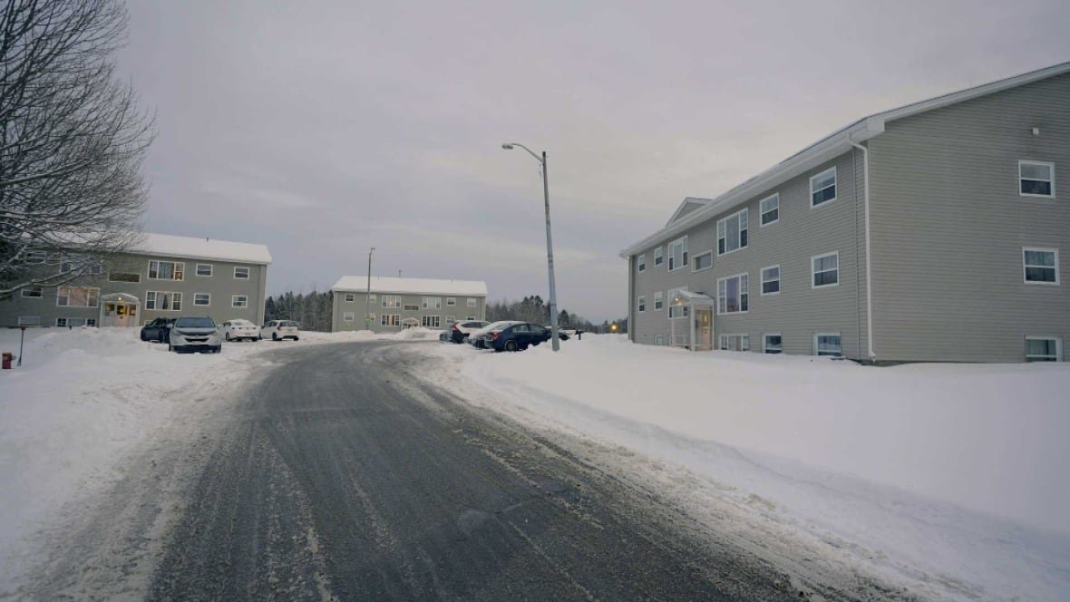 A snow-lined road leading up to three low-rise apartment buildings with beige siding and several vehicles parked in front of them.