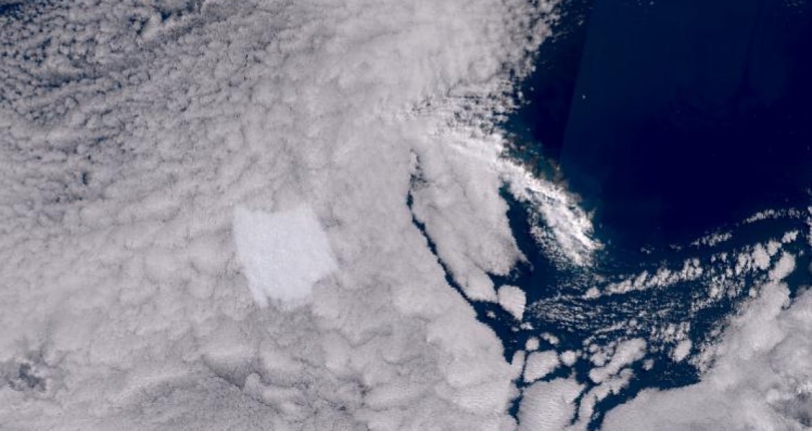 A satellite image shows a strip of an island, partially covered by clouds, and then a bright square of an iceberg near it, glowing up through full cloud coverage.