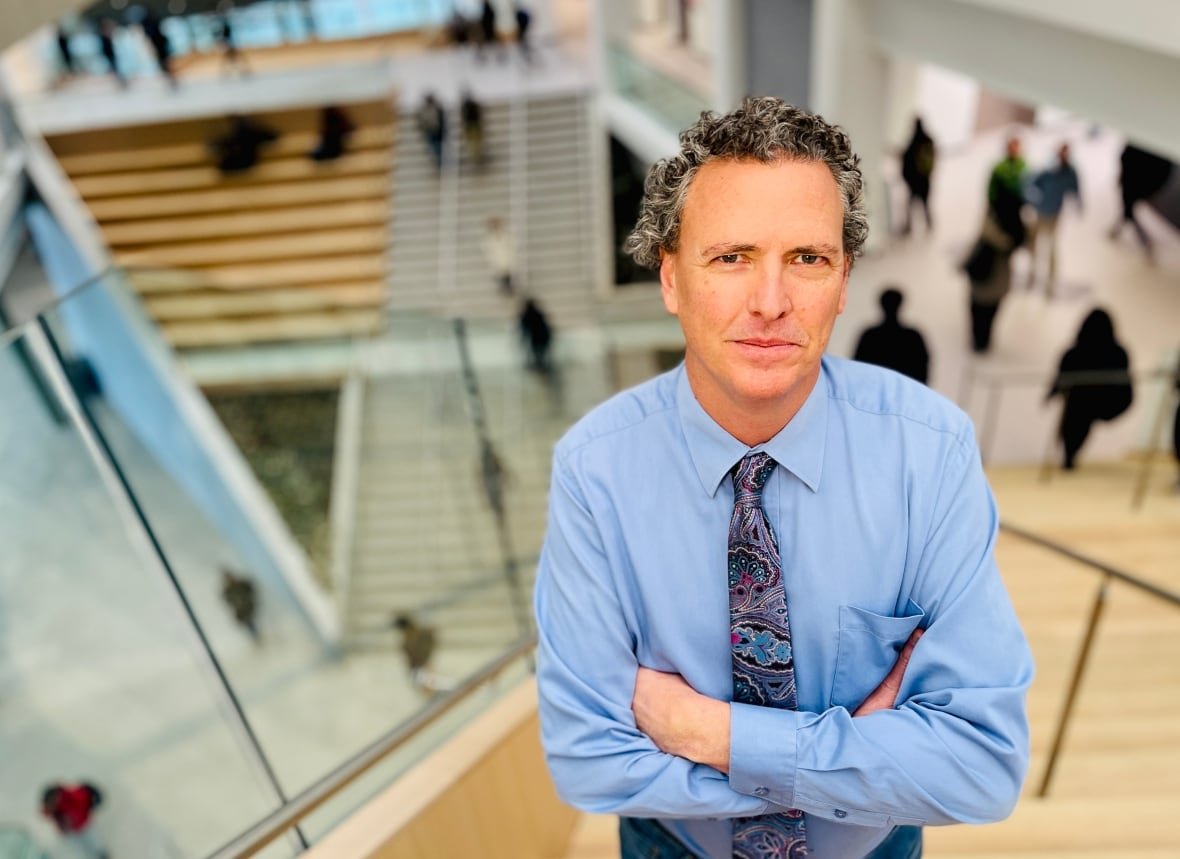 A man with curly, grey hair stands wearing a blue shirt and purple patterned tie.