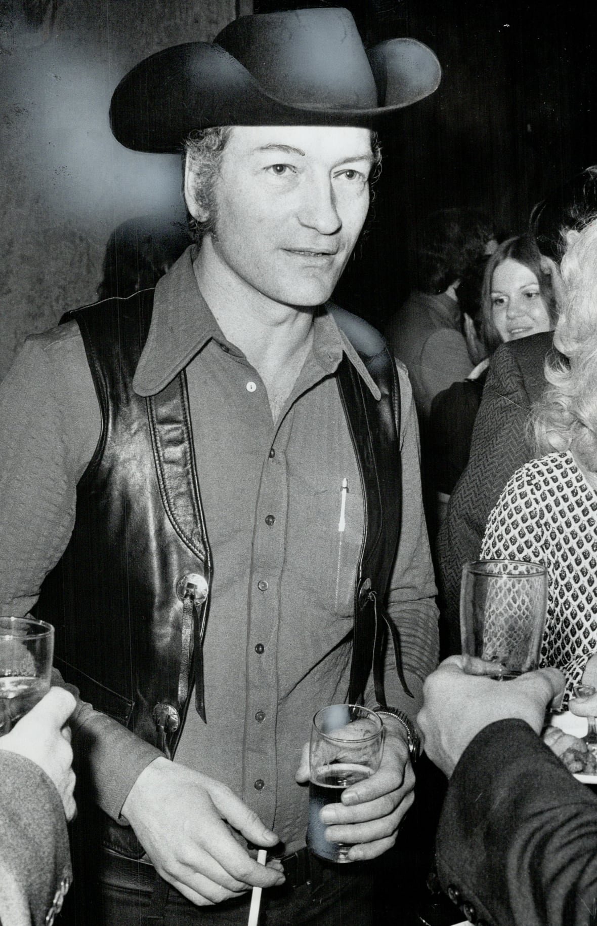 Stompin' Tom Connors at a party wearing black pants, a button up shirt and a leather vest and cowboy hat.