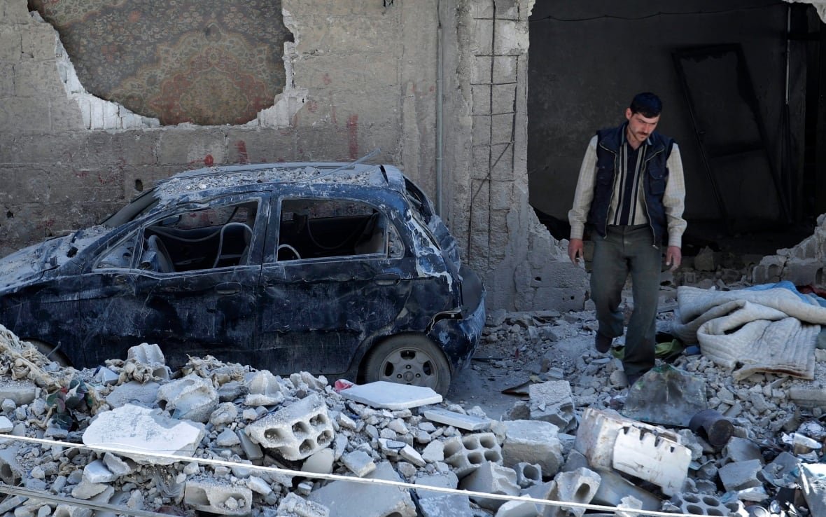 A man walks among the rubble of a destroyed building, with a car nearby.