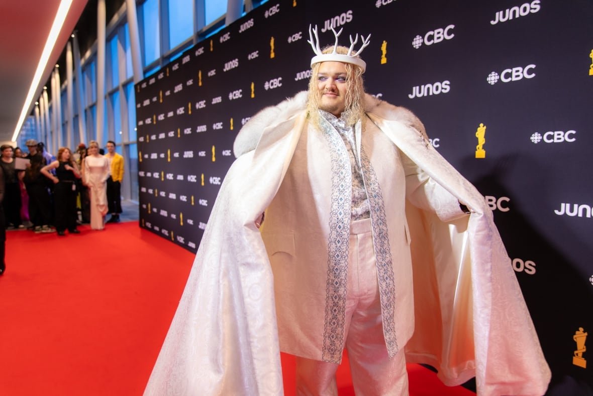 Talk on the carpet wearing a white suit with a white, fur-trimmed cape and a white antler-shaped crown.