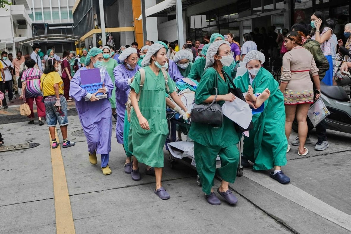 Several people wearing hospital gowns and head coverings, mostly women, are shown outside a building, wheeling an unseen patient on a gurney.