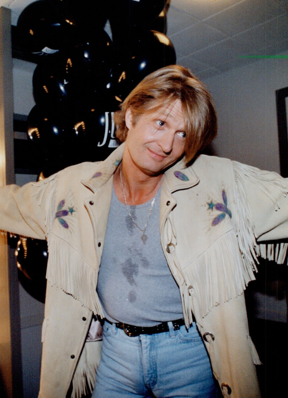 Tom Cochrane wearing blue jeans, a grey T-shirt and a beige, fringed leather jacket.