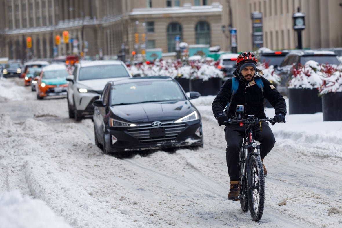A cyclist leads a line of cars down Front Street in front of Union Station in Downtown Toronto during the day time. The road and all surfaces are covered in snow