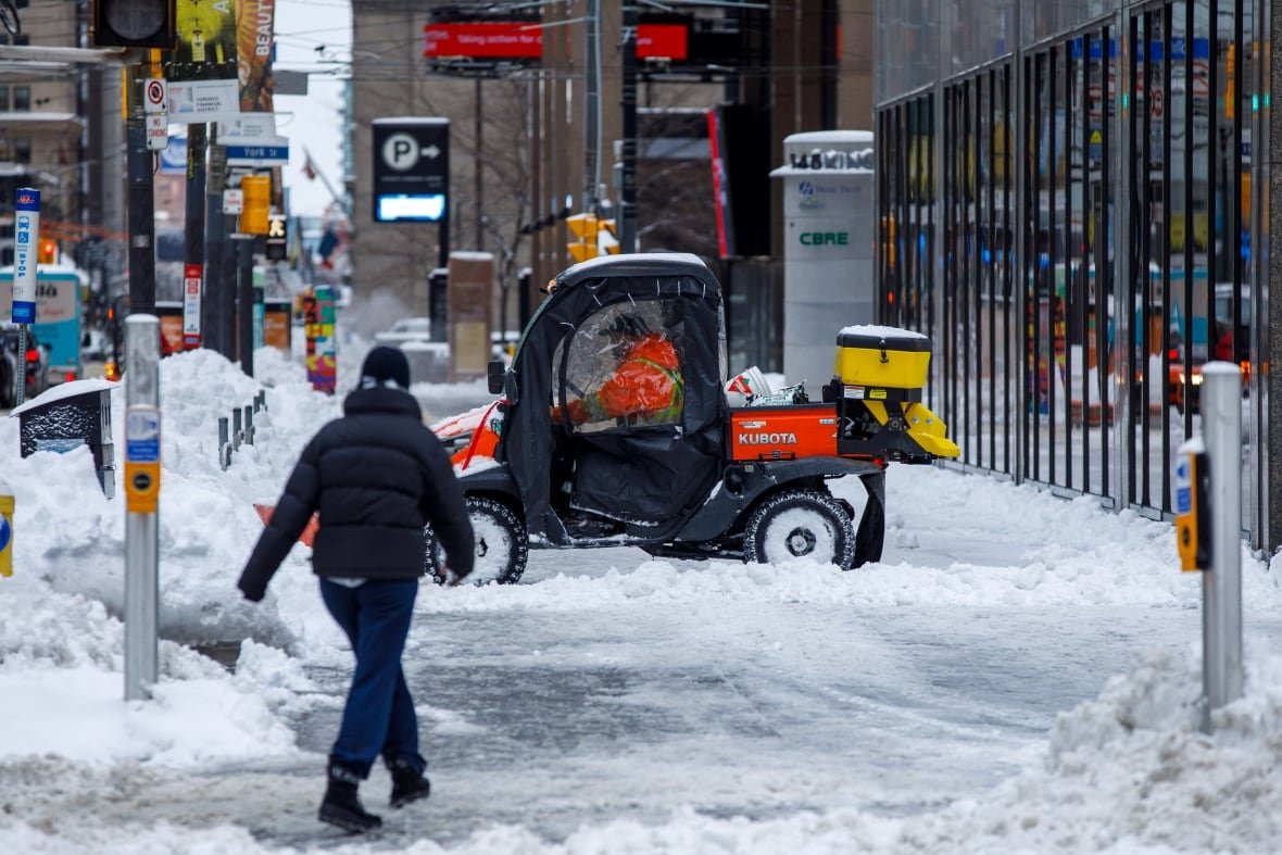 toronto-snow-cleanup-4 A city worker uses a small plow to clear a sidewalk in downtown Toronto of snow. Piles of snow surround the sidewalk as a pedestrian passes.