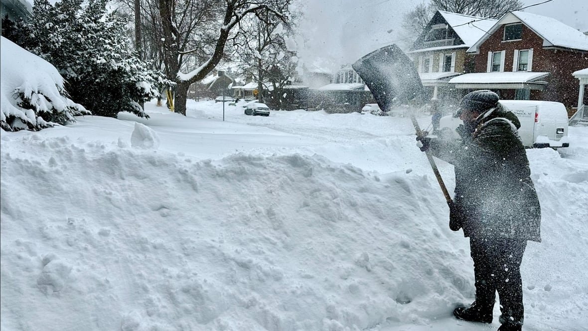 A man in winter gear shovels his driveway next to a 5-foot tall snow bank on a residential street covered in snow
